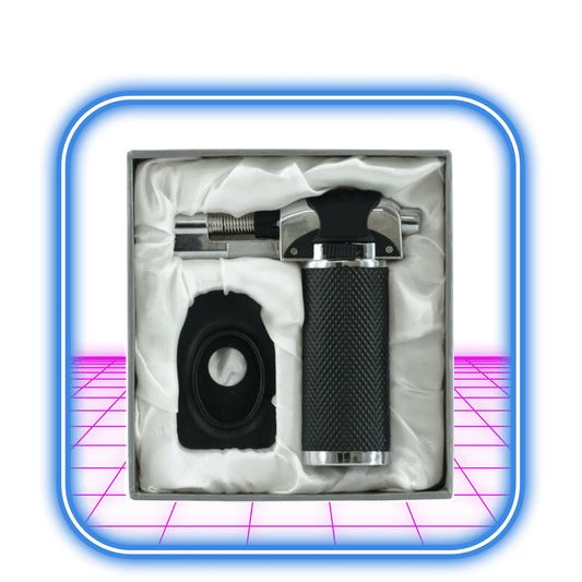 Atomic | Butan Torche Black EVP från Atomic har en svart och silver ficklampa med justerbar låga och texturerat grepp, som visas bredvid ett svart cirkulärt tillbehör i en vit vadderad fyrkantig låda med blå och rosa kanter.