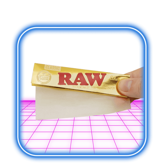 En hand håller ett paket RAW | Ethereal Classic K.S Slim rullpapper, med ett tunt ark utdraget. Bakgrunden har en blå kant och ett rosa område som liknar ett tangentbord.