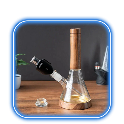 XMAX | Riggo - Svart, en vattenpipa av glas och trä med en svart skål, vilar på en träyta bredvid en e-nagel och glasbehållare, med en suddig bakgrund med växter och andra föremål.